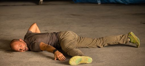 Mann liegt ausgestreckt auf dem Boden mit einem Arm über dem Kopf und trägt ein T-Shirt, Hose und Sportschuhe