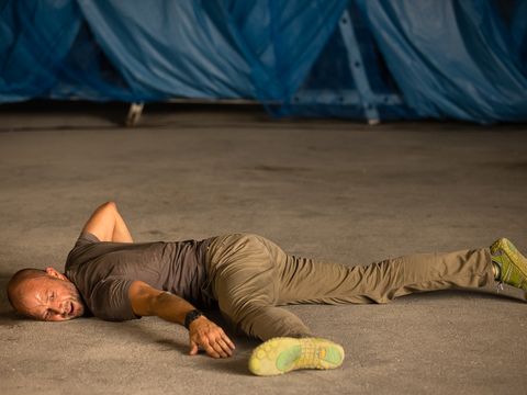 Man lying stretched out on the ground with one arm over his head wearing a t-shirt, pants, and athletic shoes