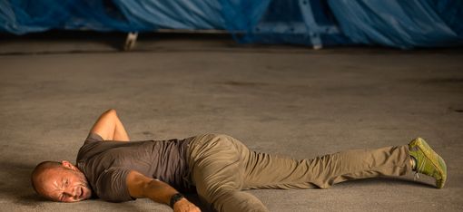 Man lying stretched out on the ground with one arm over his head wearing a t-shirt, pants, and athletic shoes