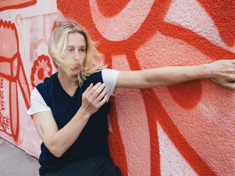 Frau mit blonden Haaren steht vor einer rot-weißen Wand mit Graffiti und streckt einen Arm aus, während ihr Haar ins Gesicht weht