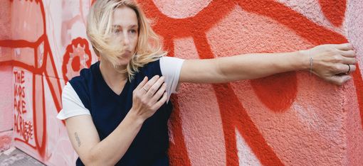 Frau mit blonden Haaren steht vor einer rot-weißen Wand mit Graffiti und streckt einen Arm aus, während ihr Haar ins Gesicht weht