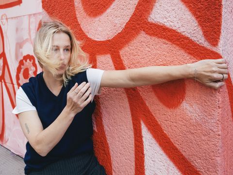Frau mit blonden Haaren steht vor einer rot-weißen Wand mit Graffiti und streckt einen Arm aus, während ihr Haar ins Gesicht weht
