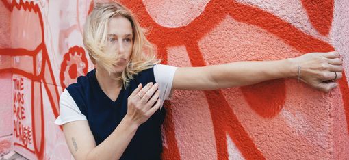 Blonde woman standing against a red and white graffiti wall with one arm extended and hair blowing across her face