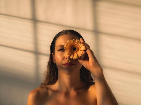 Frau mit nackten Schultern hält eine Blume vor ihr rechtes Auge vor einem hellen Hintergrund mit Schattenlinien