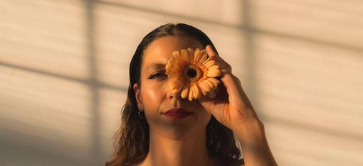 Woman with bare shoulders holding a flower in front of her right eye against a light background with shadow lines