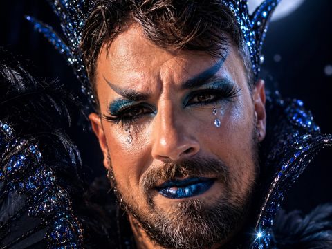 Mann mit Bart und auffälligem blau-schwarzem Make-up, trägt eine mit Edelsteinen besetzte Krone und ein schwarzes, federverziertes Kostüm vor einem Vollmondhintergrund