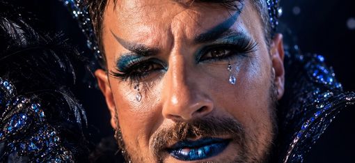 Mann mit Bart und auffälligem blau-schwarzem Make-up, trägt eine mit Edelsteinen besetzte Krone und ein schwarzes, federverziertes Kostüm vor einem Vollmondhintergrund