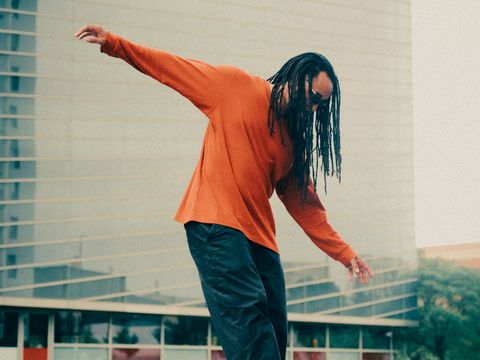Person mit langen Dreadlocks und orangefarbenem Pullover balanciert auf einer Mauer vor einem modernen Gebäude