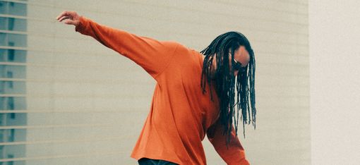 Person mit langen Dreadlocks und orangefarbenem Pullover balanciert auf einer Mauer vor einem modernen Gebäude