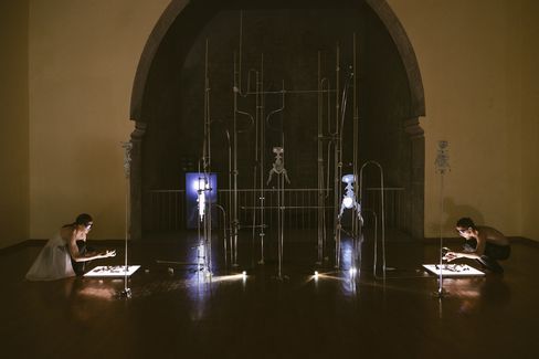 Two people kneel on opposite sides of illuminated surfaces with metal rods and apparatuses in a dark room with a large arch in the background.