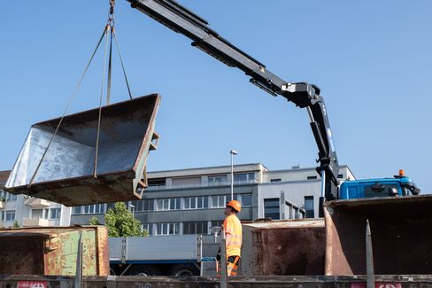 Construction worker in orange safety clothing and helmet watches a crane lifting a large metal container in front of a multi-story building.