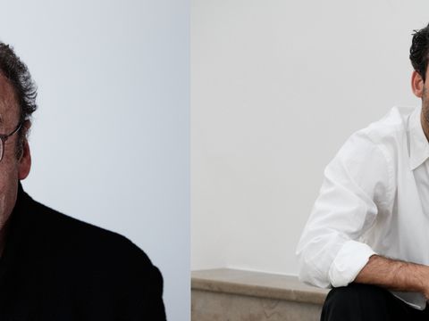 Two men with dark hair sitting against a light background, the left wearing a black shirt and the right wearing a white shirt.