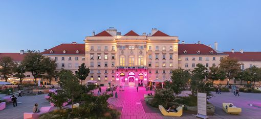 Großes historisches Gebäude mit rotem Dach und beleuchteter Fassade vor blauem Himmel, umgeben von gepflastertem Platz mit Bänken und Bäumen