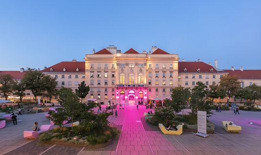 Großes historisches Gebäude mit rotem Dach und beleuchteter Fassade vor blauem Himmel, umgeben von gepflastertem Platz mit Bänken und Bäumen