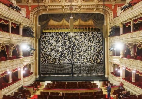 View inside a historic theater with ornate interior and a large stage featuring a curtain covered in a complex white line pattern and text.