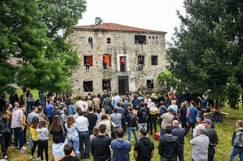 Crowd of people in front of an old stone house with several individuals looking out from windows and doors.