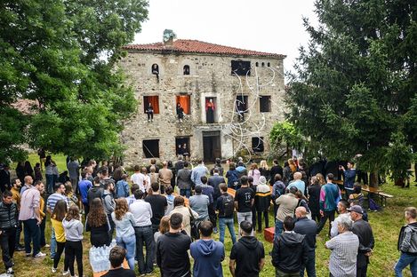 Crowd of people in front of an old stone house with several individuals looking out from windows and doors.