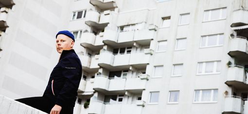 Person wearing a blue cap and dark jacket leaning on a concrete wall in front of a multi-story residential building with balconies.