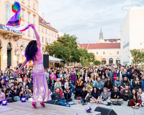 Person in langem Kleid und hohen Schuhen hebt eine Regenbogenfahne vor einer großen Menschenmenge auf einem Platz mit historischen Gebäuden.