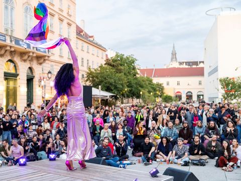 Person in long dress and high heels waving a rainbow flag in front of a large crowd on a square with historic buildings.