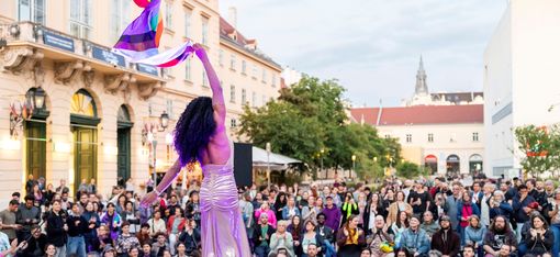 Person in long dress and high heels waving a rainbow flag in front of a large crowd on a square with historic buildings.