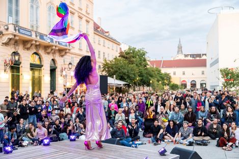 Person in langem Kleid und hohen Schuhen hebt eine Regenbogenfahne vor einer großen Menschenmenge auf einem Platz mit historischen Gebäuden.