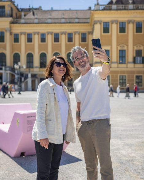 Zwei Besucher machen ein Selfie vor Schloss Schönbrunn neben einem rosa Enzi-Sitzmöbel.