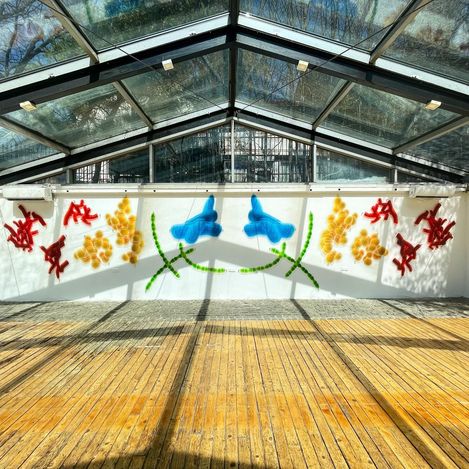 Large white wall with colorful abstract painting of red, yellow, blue, and green shapes in a room with glass roof and wooden floor.