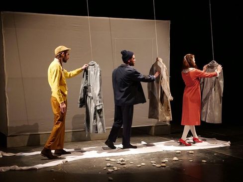 Three people on a stage each touching a hanging coat in front of a plain backdrop.