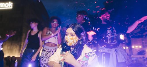 Group of people at a nighttime event with DJ booth and colorful lighting effects.
