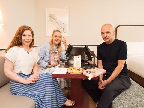 Three people sit around a small table with microphones, books, and fruit in a bright room with a bed in the background