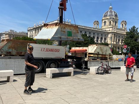 Two men stand on a paved square in front of a large rusty metal object being lifted by a crane with a historic building with a dome in the background.