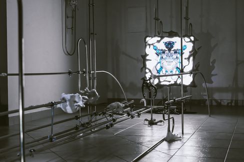 Art installation with metallic rods and a lit frame-like object on a wall featuring a blue mechanical owl motif.