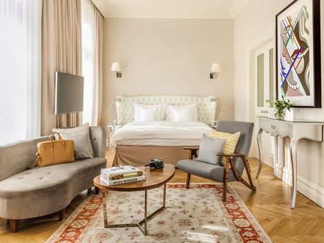 Modernes Hotelzimmer mit einem beigen Polsterbett mit weißer Bettwäsche, einer gemütlichen Sitzecke mit einem geschwungenen grauen Sofa und einem grauen Sessel, einem runden Couchtisch aus Holz, einem dekorativen Teppich, einem kleinen Konsolentisch und einem an der Wand montierten Flachbildfernseher; große Fenster mit Vorhängen lassen natürliches Licht in den Raum strömen, ergänzt durch stilvolle Kunstwerke an der Wand.