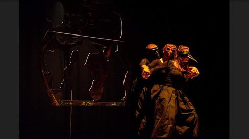 Three people in dark clothing and masks stand close together in front of a large abstract metal sculpture on a dark stage.
