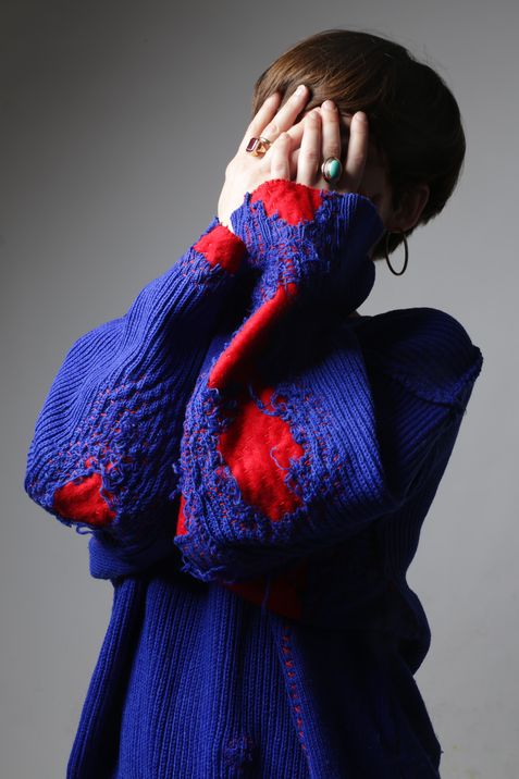 Person with short hair wearing a blue sweater with irregular red patches covering their face with hands showing rings.