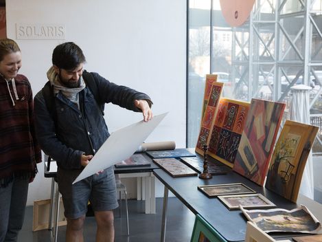 Zwei Personen betrachten ein großes Blatt Papier in einem Raum mit mehreren Bildern und Kunstwerken auf einem Tisch neben einem Fenster.