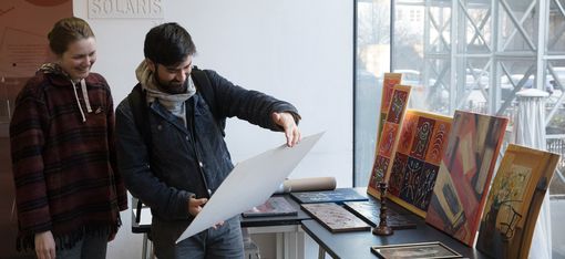 Zwei Personen betrachten ein großes Blatt Papier in einem Raum mit mehreren Bildern und Kunstwerken auf einem Tisch neben einem Fenster.