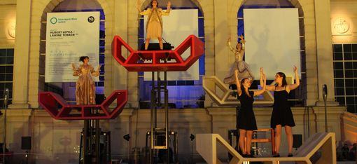 Dancers perform on colorful Enzos lifted in front of the illuminated façade of Kunsthalle Wien during a nighttime performance at MuseumsQuartier.