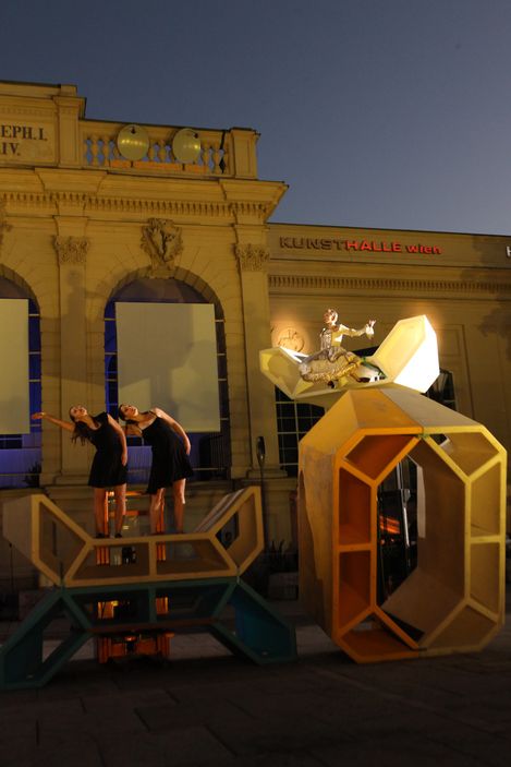 Several dancers perform on an Enzo suspended by a crane, floating in the air during a nighttime dance performance at MuseumsQuartier.