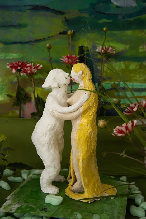 Two abstract clay figures, one white and one yellow, embracing on a green surface with red flowers and green leaves in the background.