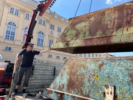 Person standing next to a crane touching a large rusty metal piece in front of a historic building.