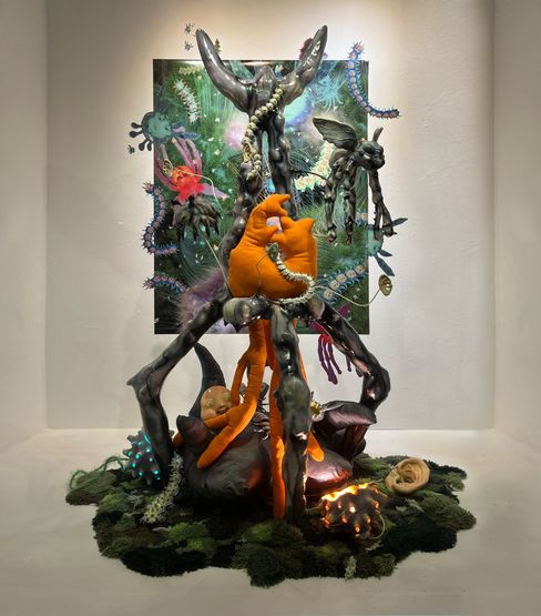 Sculpture with black organic-shaped rods, orange fabric figures, and glowing spiked objects on moss-like base in front of an abstract colorful painting.