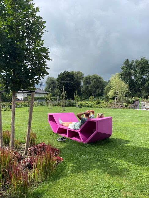 Eine Person entspannt sich auf einem leuchtend pinkfarbenen Enzi auf einem üppig grünen Rasen. Bäume und ein bewölkter Himmel umrahmen die friedliche Szene im Freien.