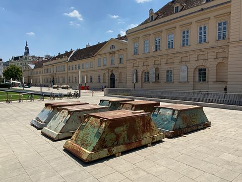 Several rusty trapezoid-shaped metal objects stand on a paved square in front of a historic building with many windows.