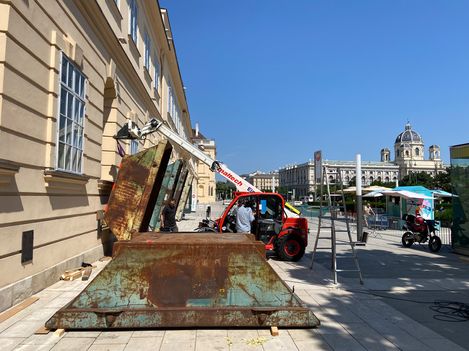 Rusty metal containers in front of a historic building with people and a red telehandler on a paved square under a clear blue sky.