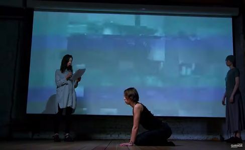 Three people on a stage in front of a large screen, one standing holding paper, one kneeling on the floor, the third standing to the side.