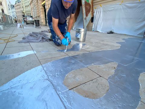 Person kneeling on a sidewalk painting different shapes on the ground with gray paint in an urban setting.