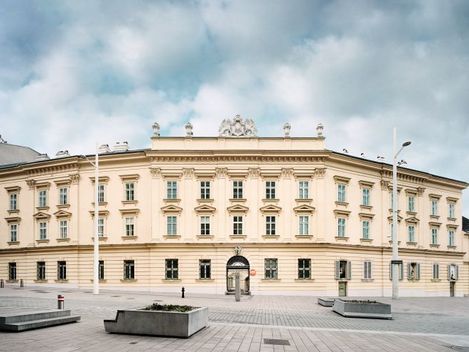 Großes klassizistisches Gebäude mit symmetrischer Fassade und mehreren Fenstern vor bewölktem Himmel