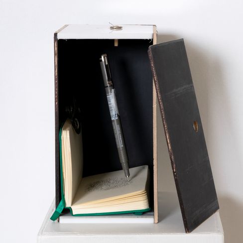 Open black box with an open notebook inside and a pen standing upright in it.
